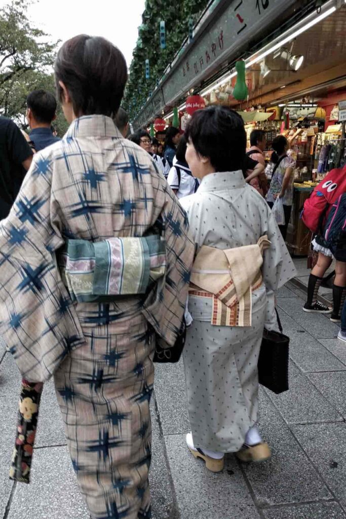 Two women in traditional kimonos walk through a busy Tokyo street market outside Sensoji Temple filled with stalls, lanterns, and souvenirs.