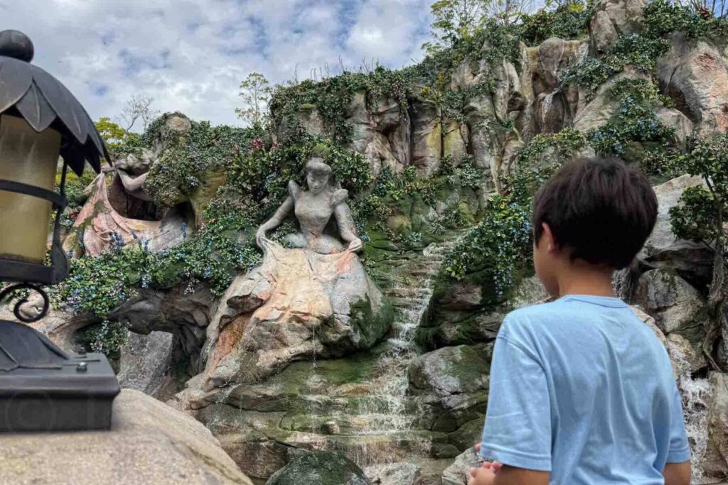 A boy in a blue shirt looks at carved princess statues and waterfalls built into a rock garden at Tokyo DisneySea in the Fantasy Springs area.