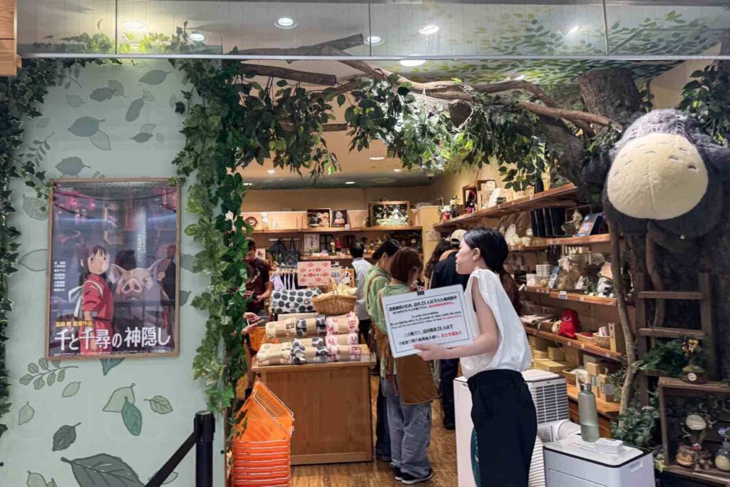 The entrance to a Studio Ghibli store in Tokyo’s Character Street with Totoro plushies, movie posters, rolled up blankets and lush forest-themed decor. A shop worker stands at the entrance with a sign in Japanese indicating the max guests allowed inside at one time.
