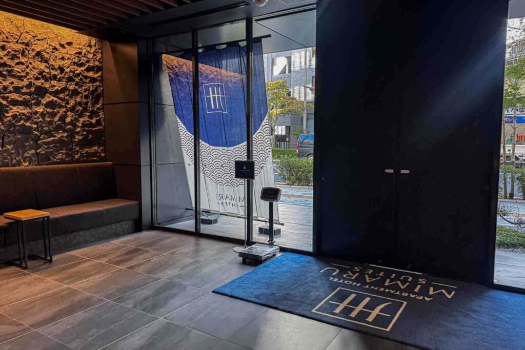 Entryway of Mimaru Suites Tokyo Asakusa family apartment hotel showing a dark stone-accent wall, seating bench, and noren curtain at the glass door. 