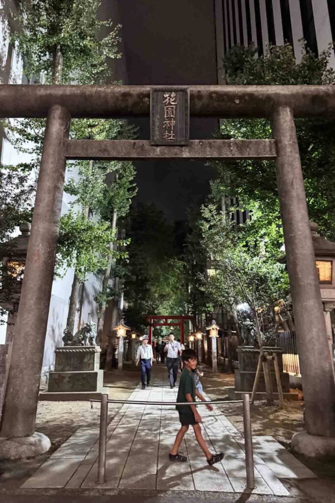 A modern city backdrop with skyscrapers on either side frames a large gray torii gate at a shrine entrance in central Tokyo. A child is walking under the middle.