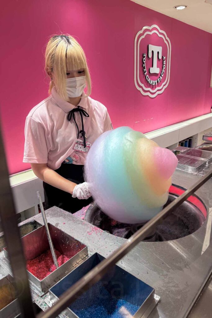 A staff member wearing a mask at Totti Candy Factory in Harajuku spins a massive pastel rainbow cotton candy against a bright pink backdrop.