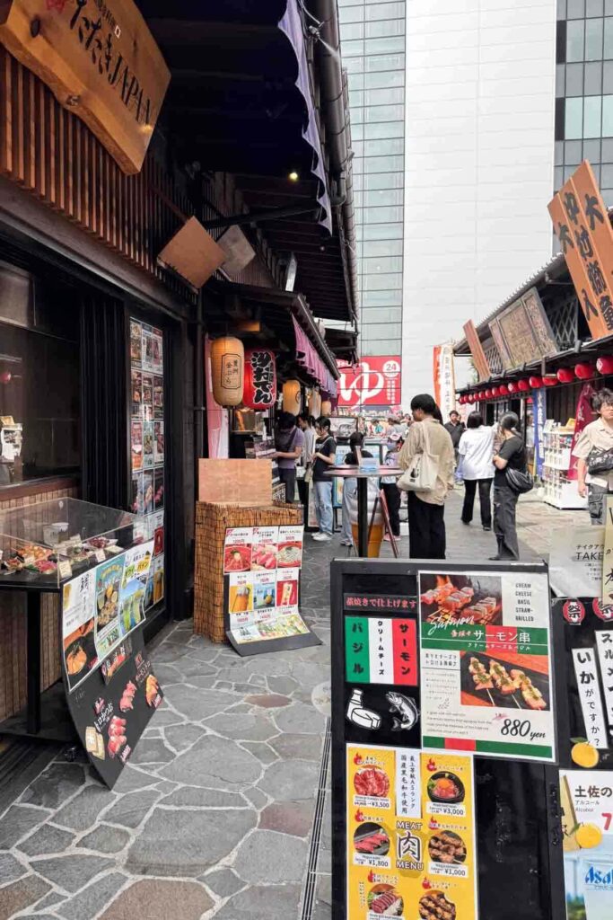 People browse food stalls in a narrow section of Tokyo’s Toyosu Market shown with lanterns, outdoor menus, and grilled skewers on display.
