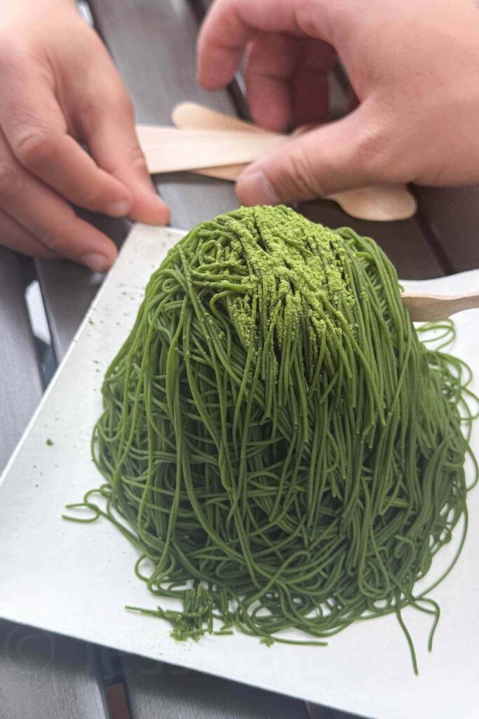 A towering matcha Mont Blanc dessert topped with powdered green tea is served on a white plate as two hands reach for wooden spoons. They dig in to enjoy the treat at Toyosu Market in Tokyo.