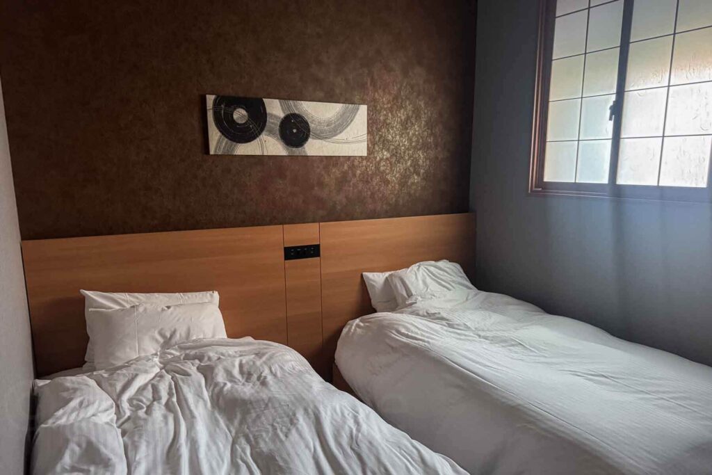Twin beds aligned side by side below a wooden headboard with white linens and a decorative brown wallpaper accent wall under soft daylight from a shoji window. Clean and comfortable bedroom setup at a family suite in Mimaru Suites Asakusa.
