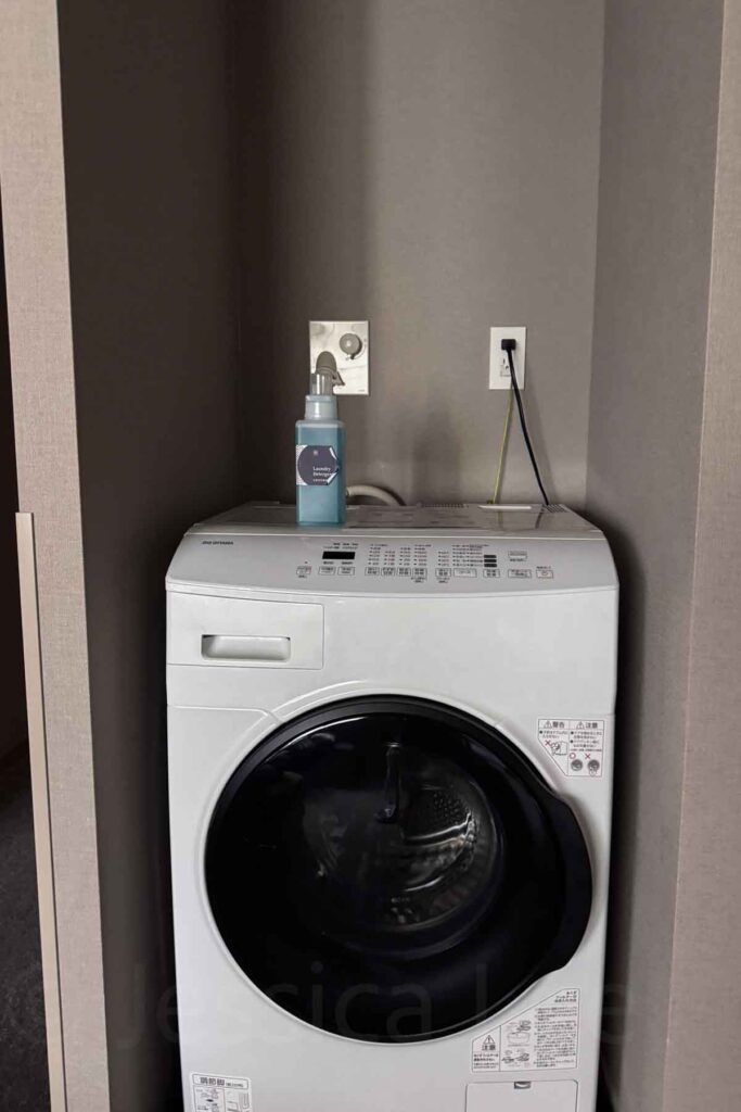 In-room white front-load washer and dryer combo with blue laundry detergent bottle on top in a Tokyo Mimaru family hotel suite. 