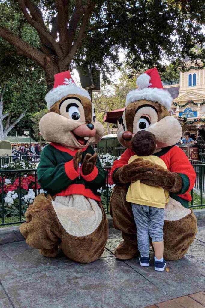 A child wearing a yellow sweatshirt and gray pants hugs Chip while Dale claps next to him, both wearing Santa hats and festive Christmas sweaters at Disneyland.