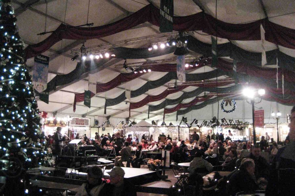 Inside a large tented Christmas market draped with red and green cloth filled with holiday lights, food vendors, and groups of people enjoying live entertainment at Christkindlmarkt in Bethlehem, Pennsylvania.