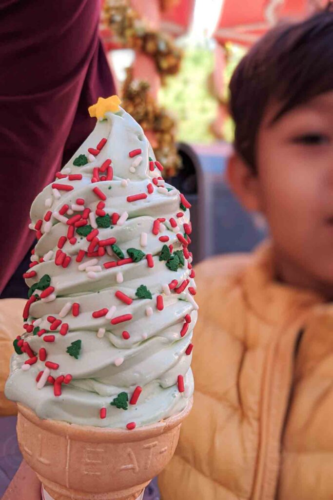 A festive soft-serve ice cream cone shaped like a Christmas tree, topped with a yellow star sprinkle and red, white, and green sprinkles covering the light green mint swirled ice cream, held by a child in a yellow jacket. A perfect treat for kids at one of the most magical Christmas destinations.