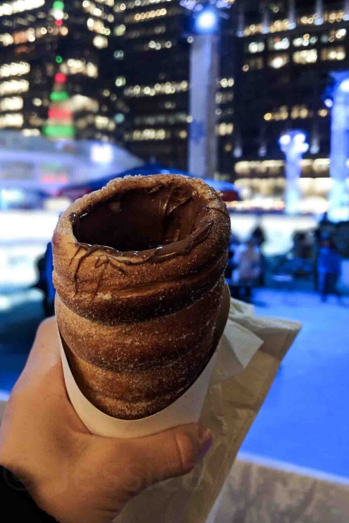 A hand holds a warm chimney cake covered in sugar and filled with Nutella, with sparkling city lights and a skating rink in the background at Bryant Park in NYC during winter.