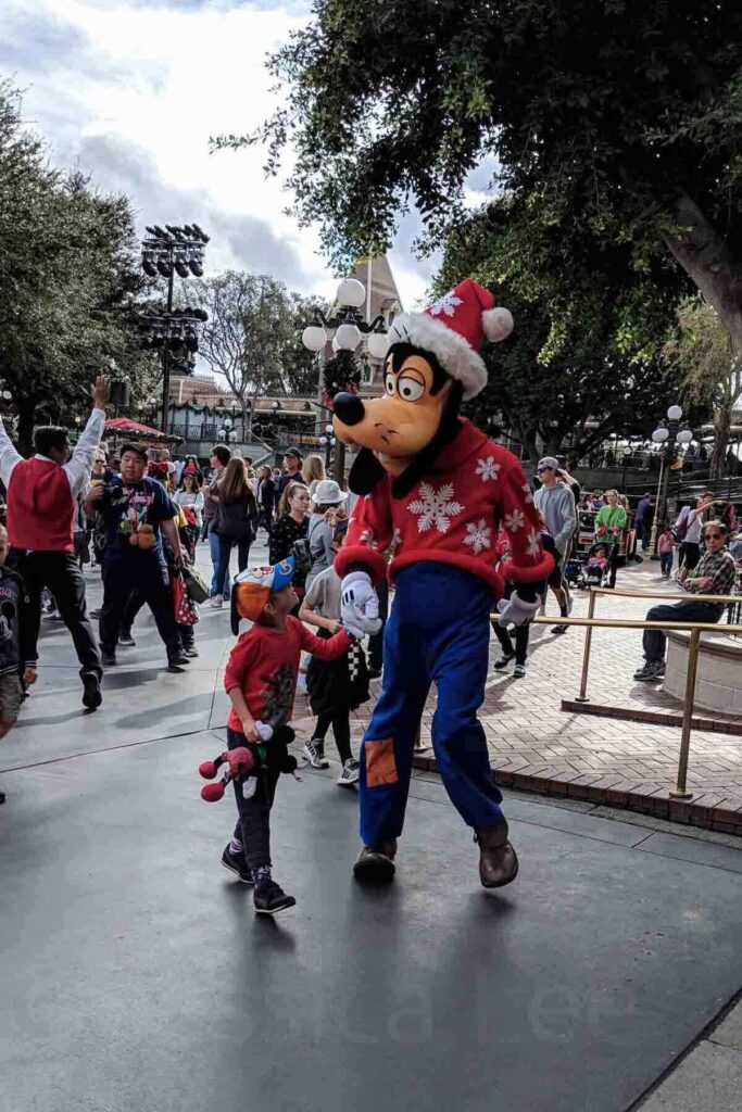 A child dressed in red shakes hands with Goofy in a Santa hat and red sweater with snowflakes at Disneyland during the holidays. A fun‑filled stop for families looking for Christmas destinations with kids.