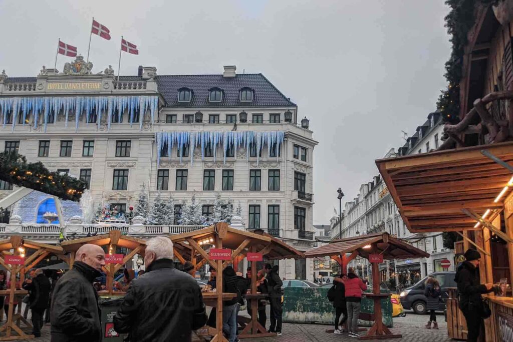 A Christmas market in Copenhagen with cozy wooden stalls and Danish flags waving atop the grand Hotel D’Angleterre, adorned with giant icicle decorations. A magical city winter scene perfect for a Christmas holiday trip.