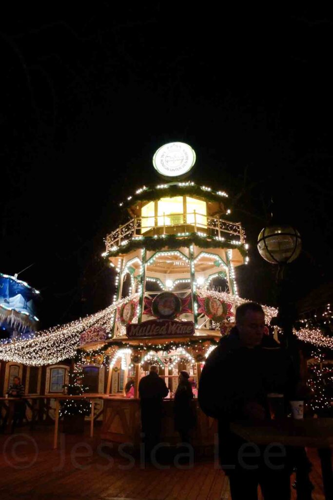A warmly lit tower‑style mulled wine stall at night wrapped in Christmas lights, with people enjoying drinks below in London during the holidays.