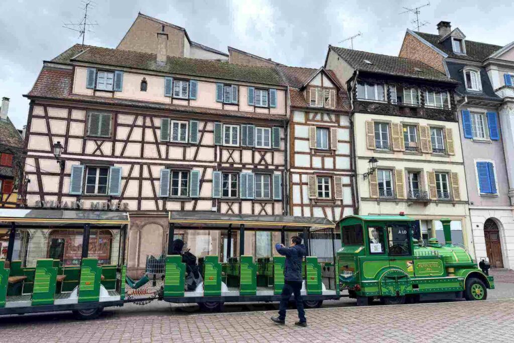 A green tourist train parked in front of colorful half‑timbered buildings in Colmar, France. A visitor takes photos.