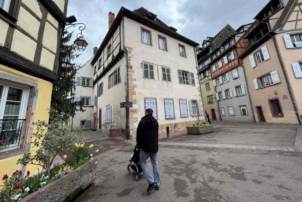 A man pushes a Yoyo stroller through an open courtyard in charming Colmar, France lined with pastel half‑timbered buildings and flower beds.