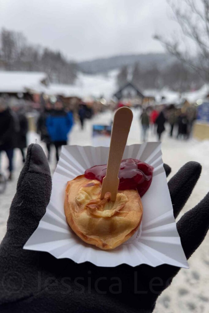 A hand in a black glove holds a warm oscypek fried cheese snack with red berry sauce on a paper tray, with a snowy European‑style Christmas market blurred in the background in Zakopane, Poland.