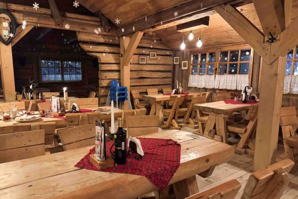 A rustic wooden lodge dining room with wood walls, beams, tables and wooden benches, festive red tablecloths, and snowflake decorations hanging from the beams. A warm and welcoming spot for traditional Polish food in Zakopane.