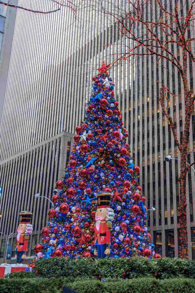 A tall Christmas tree covered in red, blue, and gold ornaments stands in front of a high-rise office building, flanked by two large toy soldier nutcrackers and wrapped in festive lights in New York City.