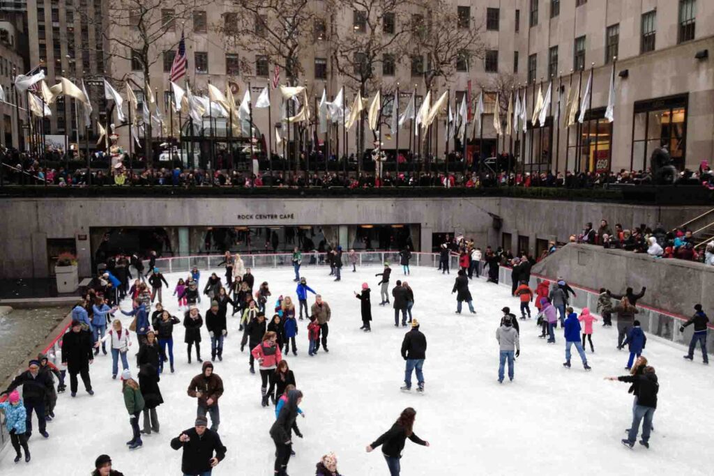 Crowds of people skate at the iconic Rockefeller Center ice rink in New York City, surrounded by tall buildings and silver and gold holiday flags. One of the most magical Christmas destinations for festive city fun.