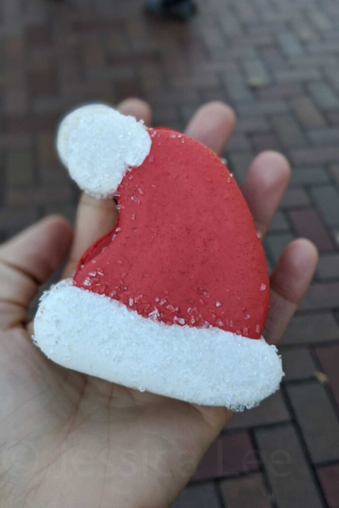 A hand holds a red and white Santa hat-shaped macaron cookie coated in sugar crystals. A sweet holiday treat picked up at Disneyland California.