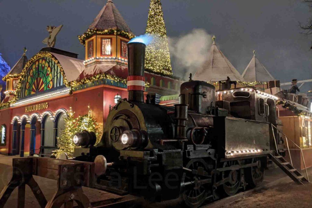 A black vintage steam train sits decorated for the holidays with lights and garlands, next to a whimsical red building labeled “HULKØBING” and a lit Christmas tree under a twilight sky inside Tivoli Gardens during December in Copenhagen.