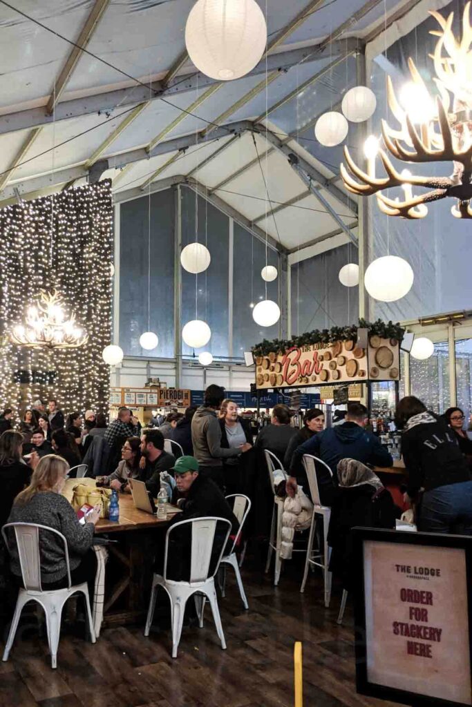 A bustling indoor holiday market “The Lodge” in NYC’s Bryant Park with hanging lanterns, twinkling string lights, and people gathered around wooden tables on white metal chairs between food stalls. A cozy retreat from the cold during a Christmas holiday trip.