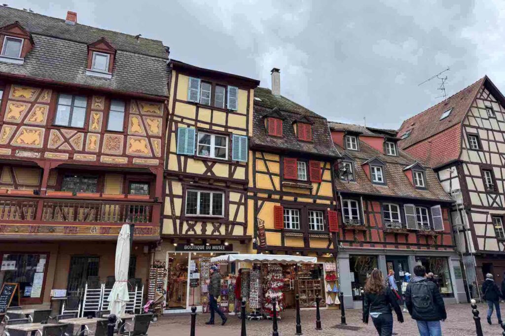 A busy pedestrian street lined with timber‑framed buildings in festive Alsatian style, in Colmar, with decorated balconies and shop signs. A magical scene in one of Europe’s most Christmassy places to visit.