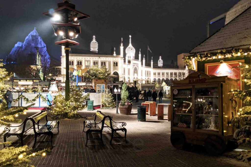 A scene during Christmastime in Tivoli Gardens in Copenhagen, buildings lines with lights, garland strung and Christmas trees all over, benches, small shops and displays for the holiday season.