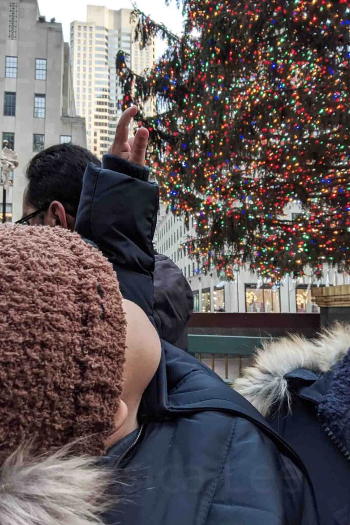A side profile of a young toddler in a brown knit hat looks up at the massive Rockefeller Christmas tree in NYC covered in multicolored lights, surrounded by tall buildings in the plaza.