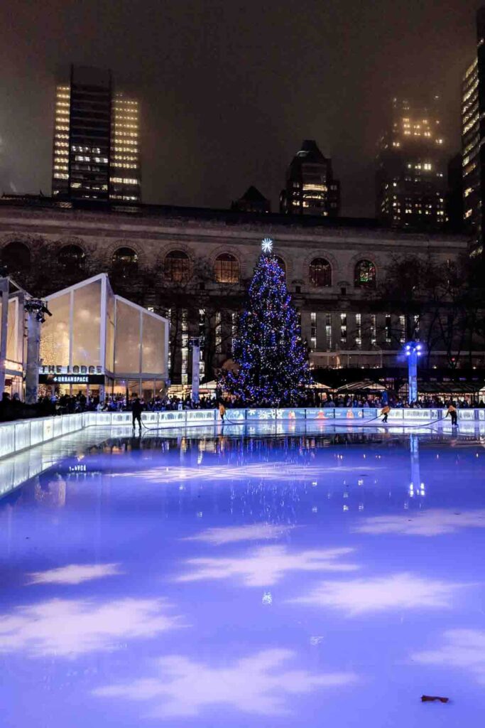 A glowing blue‑lit ice rink in front of the large decorated Christmas tree at Bryant Park in New York City, surrounded by misty skyscrapers, NYC Public Library and a urbanspace popup.