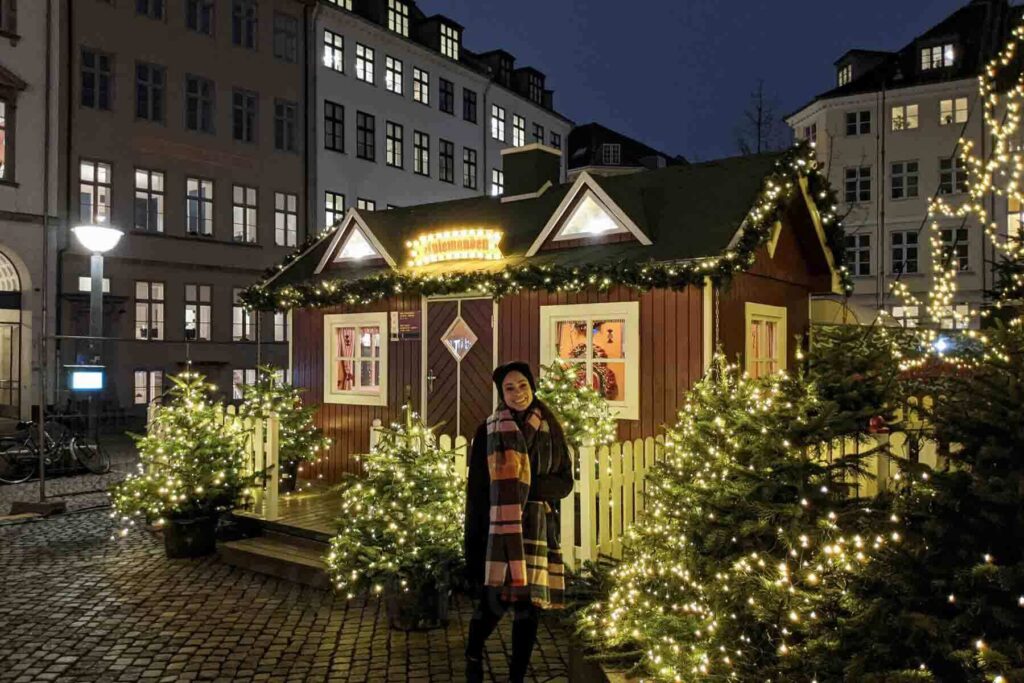 A woman wearing a colorful plaid scarf smiles in front of a wooden holiday cottage flanked by twinkling Christmas trees and lights in a small Copenhagen Christmas market at night. A fairytale Christmas destination.