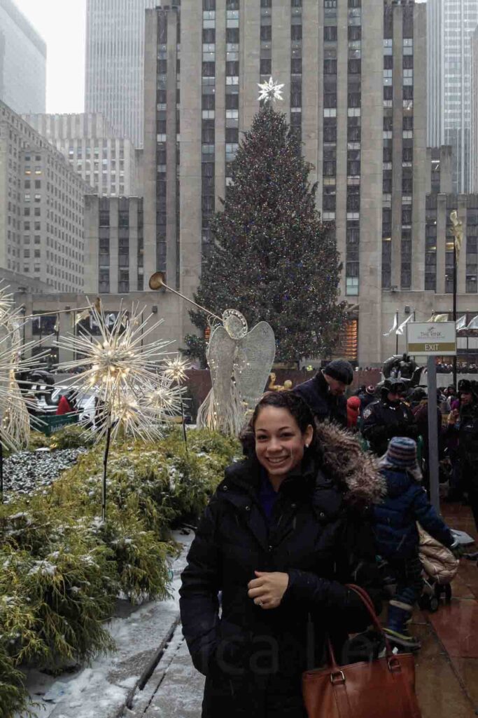 A woman smiles in front of the famous Rockefeller Center Christmas tree, surrounded by glowing angel decorations and snowy greenery. A must‑see stop during an NYC holiday trip.