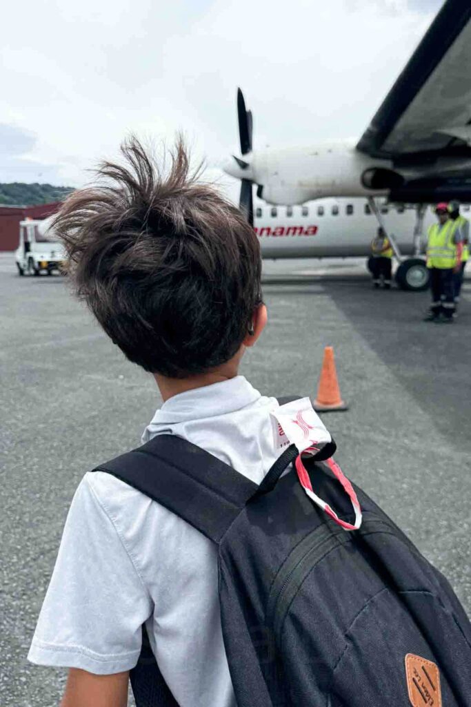 Young boy with hair blowing in the wind and a black backpack walks toward a small Air Panama airplane on the tarmac in Bocas del Toro with ground crew in yellow safety vests under the plane’s wing.