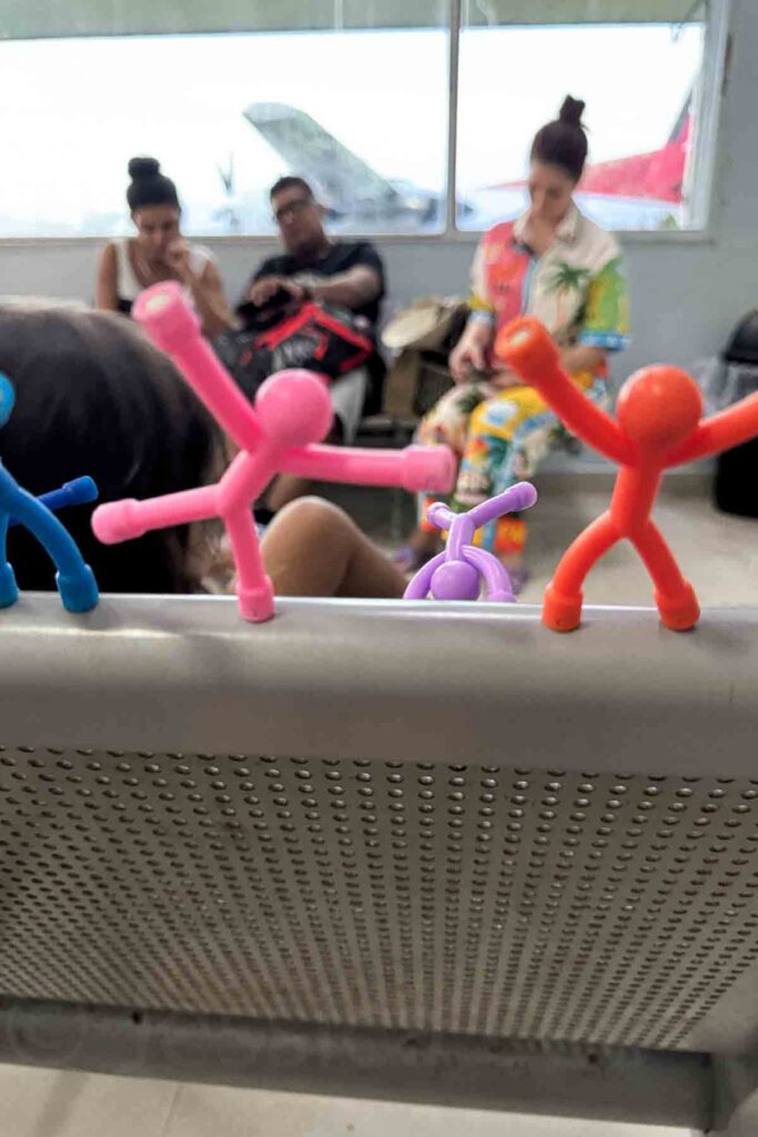 Colorful pink, blue, orange and purple stick figure toys posed on an airport bench with kids and parents waiting in the background. These travel toys for kids are great for keeping them busy before boarding.