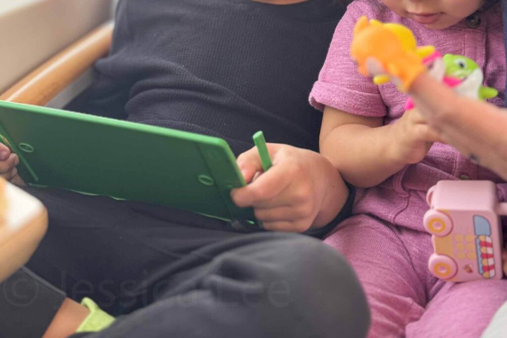 A child uses a green LCD writing tablet while a toddler in a pink jumpsuit next to them plays with small Baby Shark finger puppets and a pink rolling toy. Great example of toys to keep kids busy on a flight