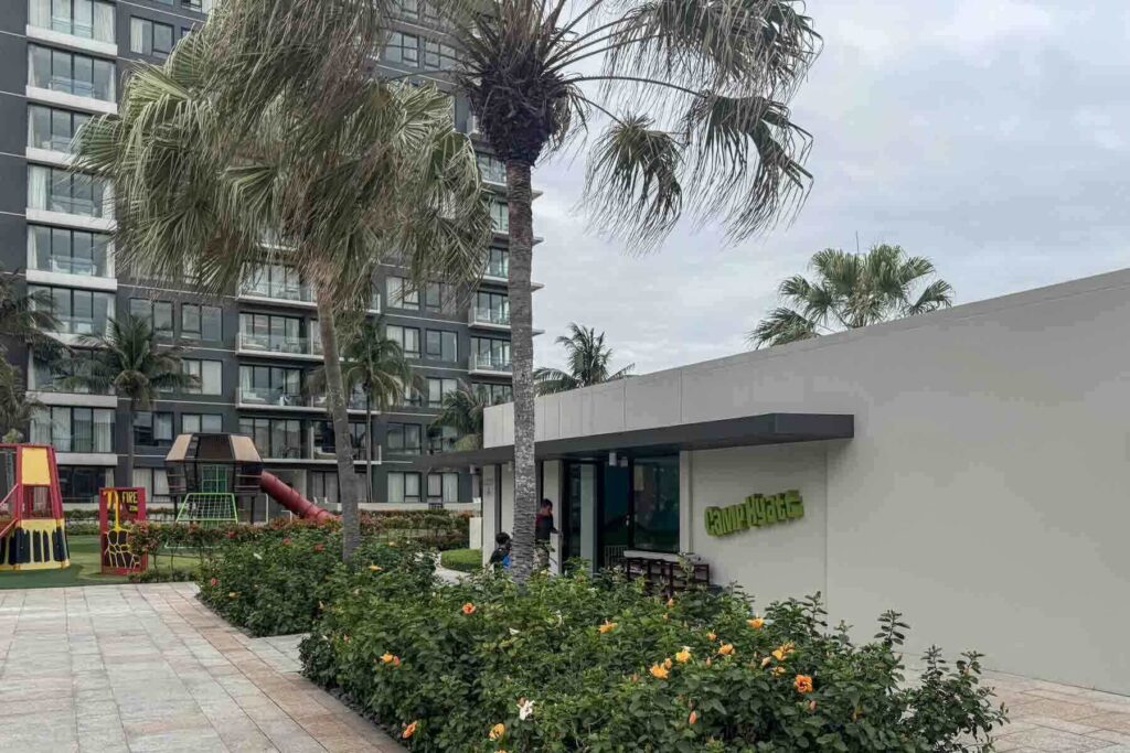 Exterior of the Camp Hyatt kids club at the Hyatt Regency Da Nang Resort, surrounded by flowers and palm trees and located beside a playground and hotel buildings.