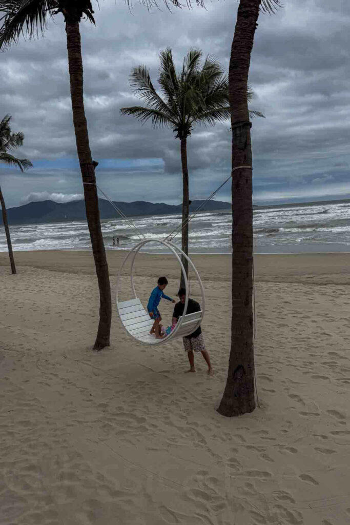 A child and adult playing on a white circular swing suspended between palm trees on a sandy beach with waves and mountains in the background at Hyatt Da Nang resort’s beach.