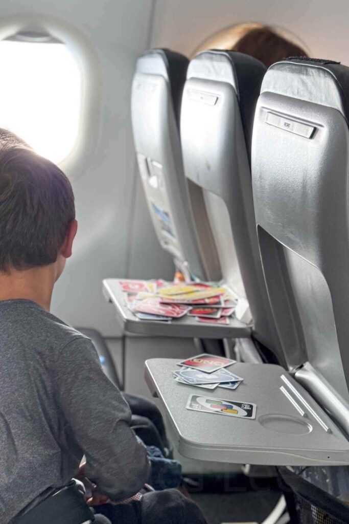 Child in a gray long sleeve shirt seated on a plane playing a game of Uno on the tray table with another child, one tray table is stacked with a bunch of cards while the other only has a few.