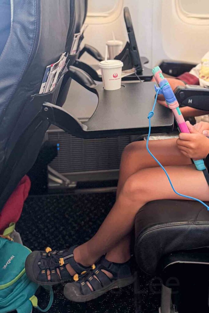 Child holding an iPad in a bright multicolored case on an airplane seat with a black tray table and white cup with a cap and straw and Japan Airlines logo on the front.
