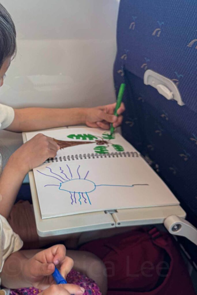 Child drawing trees and a sun in a sketchbook on an airplane tray table using markers. Check out what to pack for kids on a plane in this blog post.