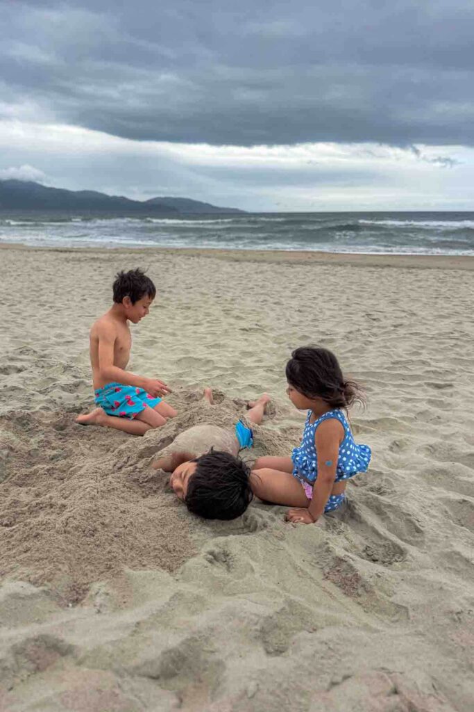Children playing on an expansive empty beach, with one child buried in the sand while two others kneel nearby with rough waves in the background of a Da Nang beach.