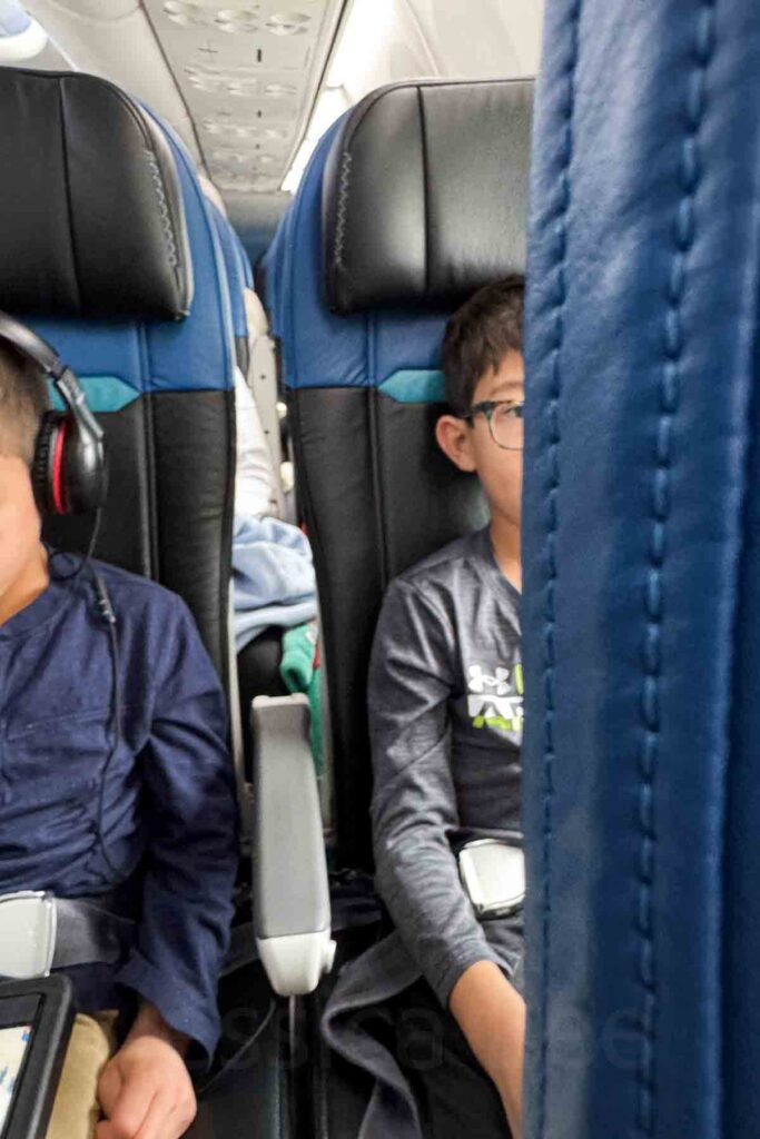 Narrow view between two black and blue airplane seats of two boys seated on a flight, one wearing headphones and watching a screen, the other wearing glasses.