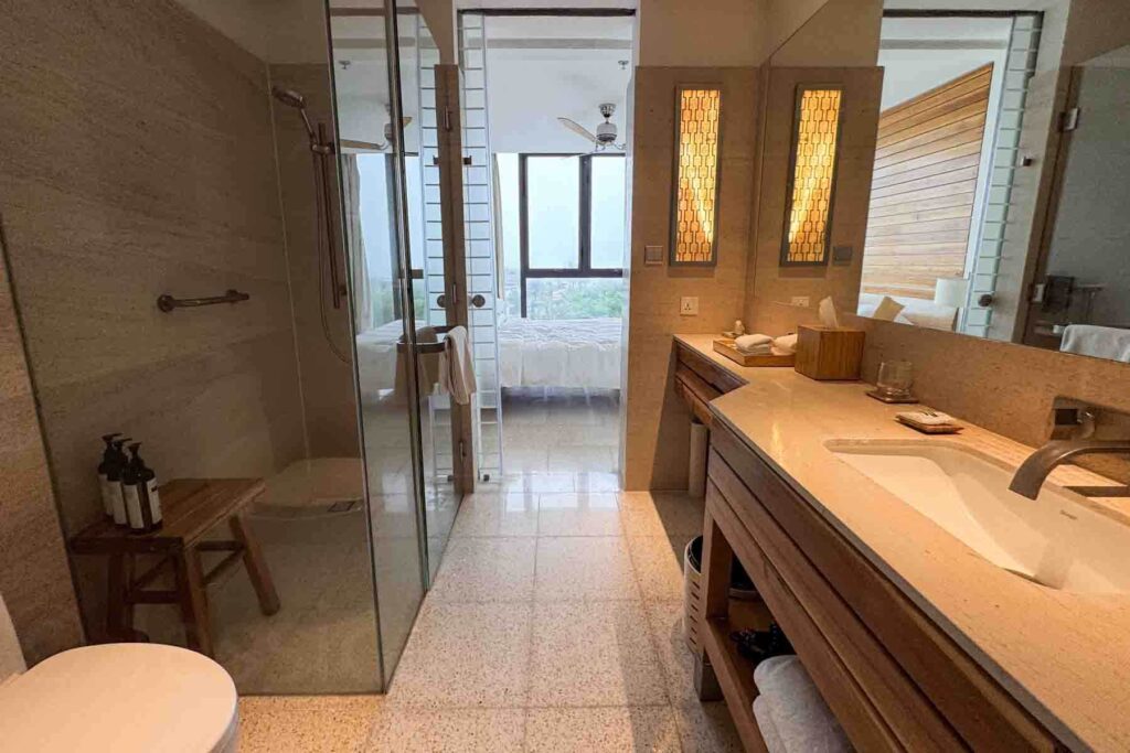 Modern ensuite hotel bathroom with an extended vanity, white towels tucked underneath, white toilet, walk-in full glass shower, and a view into the bedroom beyond at Hyatt Regency Da Nang.