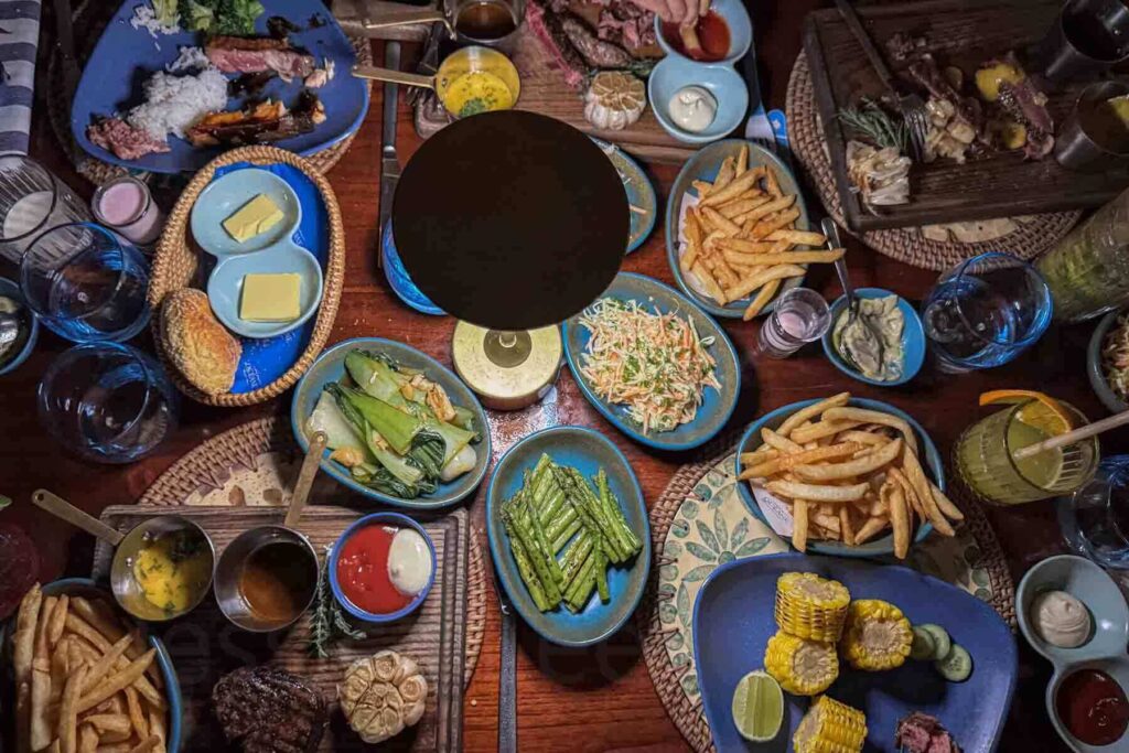 Overhead view of a large shared dinner table in the evening lit by table lamps showing off a full table of grilled meats, fries, grilled vegetables, sauces, and drinks in blue dishware served at Vive Océane Beach Club & Restaurant at Hyatt Danang Resort.
