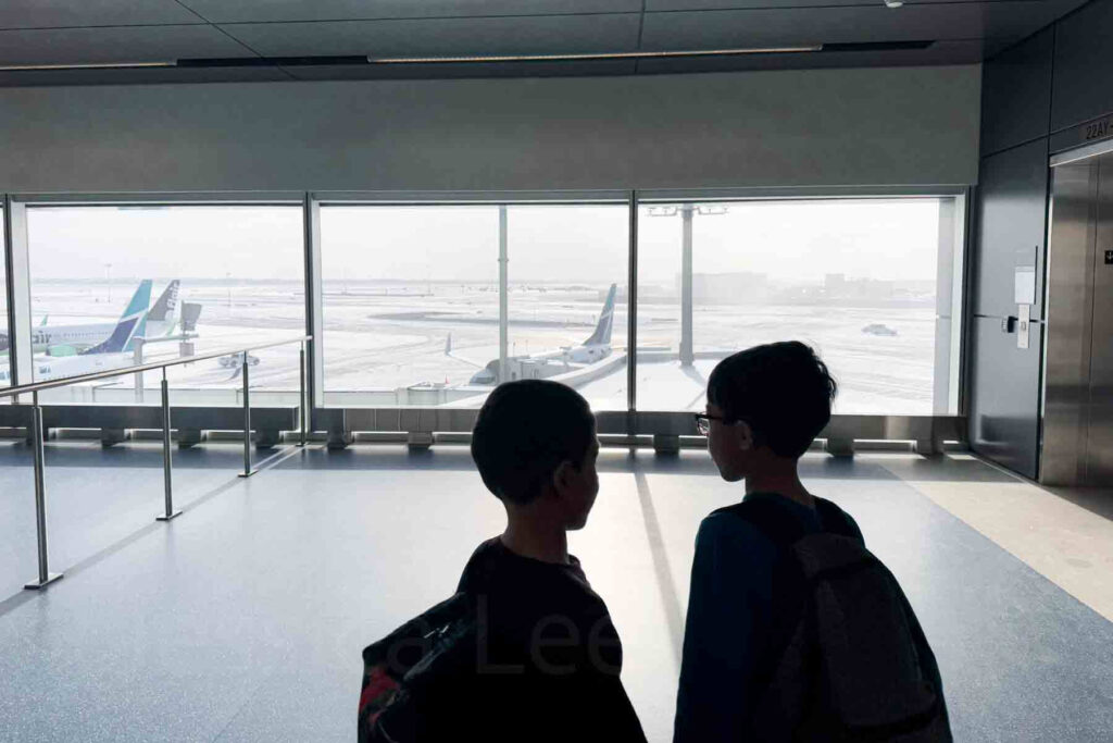 Shadowed image of two elementary-aged boys with backpacks looking out over the airport runway through large terminal windows at Calgary airport in winter with a white-out view