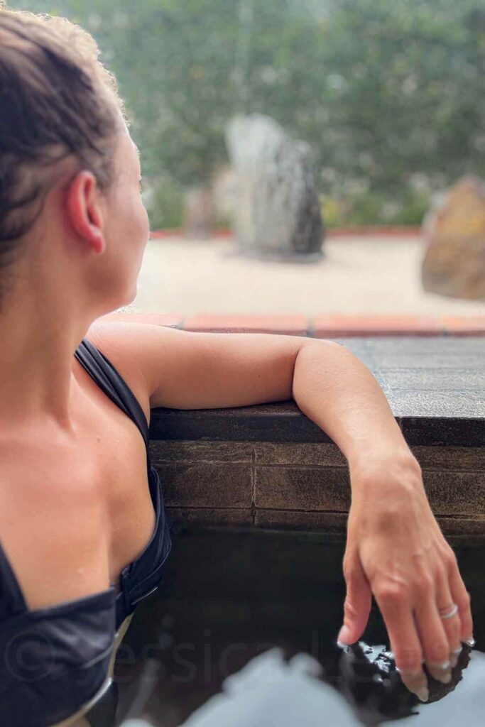 Woman in a black bikini top relaxing in a dark-tiled onsen-style hot tub at Hyatt Regency Da Nang Resort spa, gazing out at a zen Japanese garden with large stones and greenery.