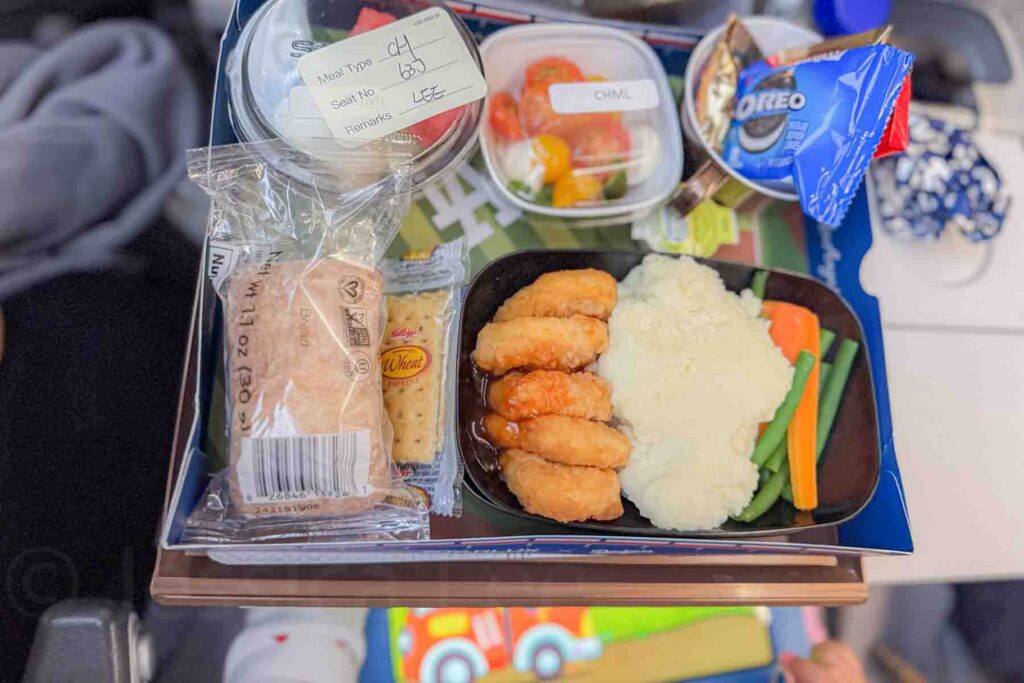 Kids airplane meal with chicken nuggets, mashed potatoes, vegetables, bread roll, crackers, fruit, and Oreo cookies served on a tray table during a long haul flight, showing what food looks like on a long flight with a toddler.