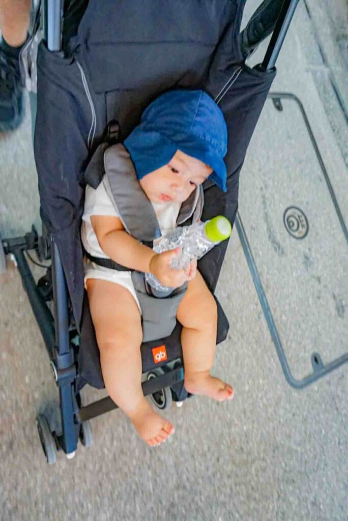 Baby wearing a blue sun hat sitting in a lightweight GB Pockit travel stroller on a city sidewalk in Japan. Demonstrates practical stroller use in Tokyo with a baby.