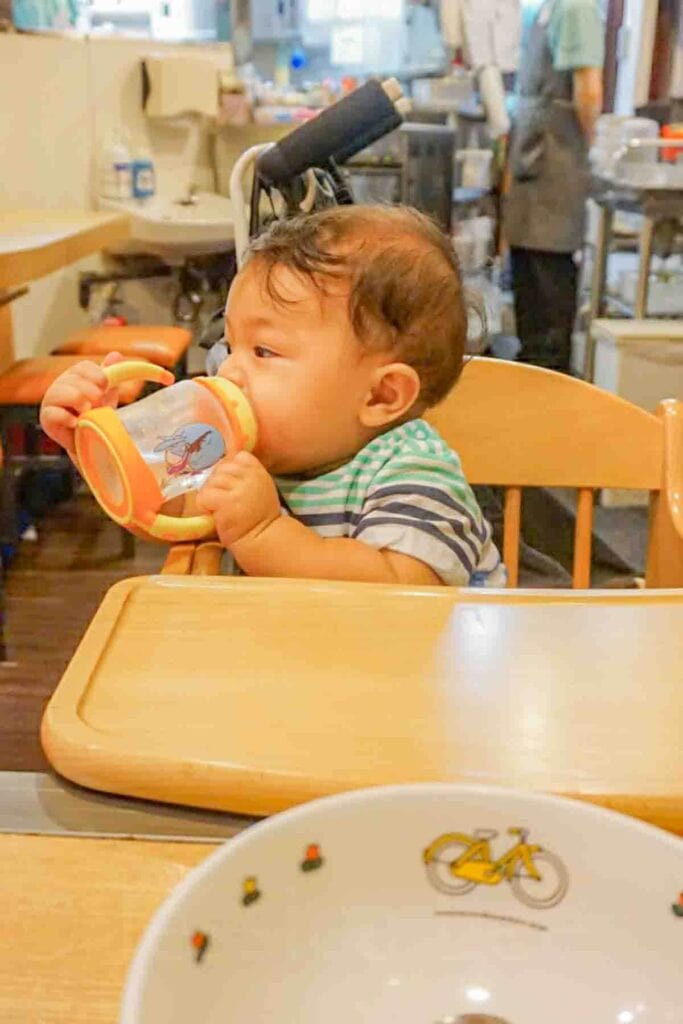 Baby sitting in a wooden high chair at a casual restaurant in Japan drinking from a sippy cup. Shows how dining out with a baby can be comfortable and convenient.