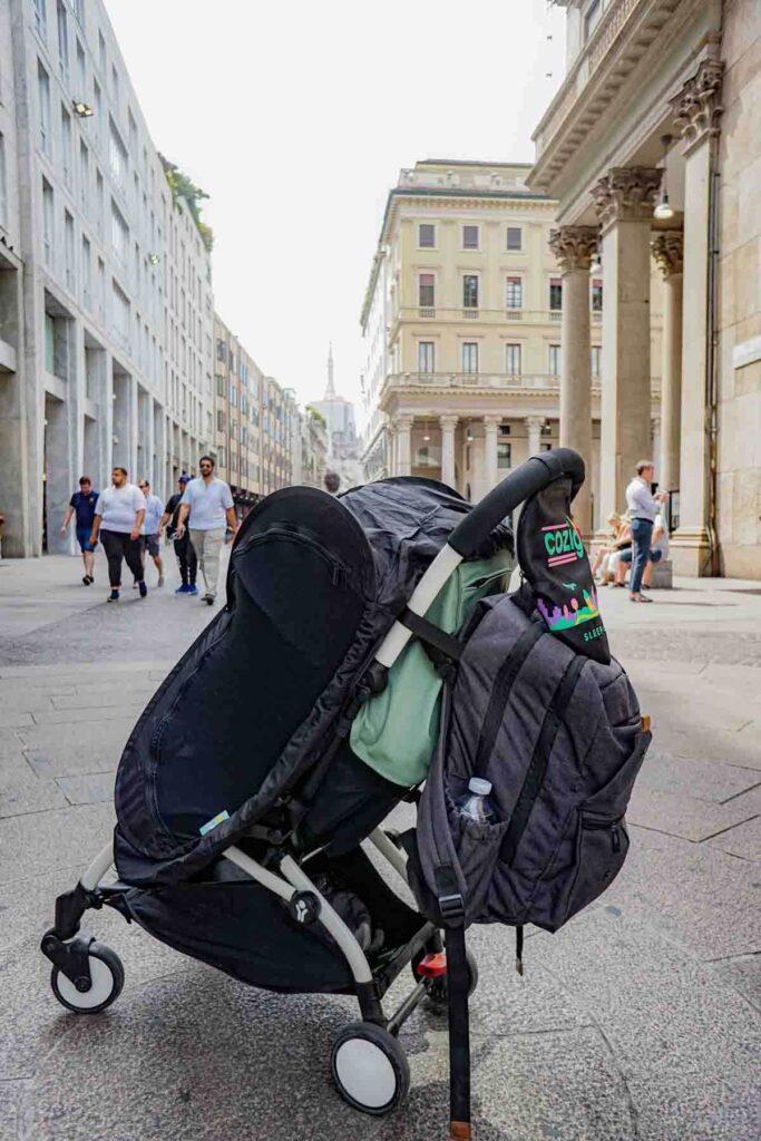 Babyzen Yoyo stroller with a blackout CoziGo sleep cover and dark gray backpack attached while walking through Milan, Italy showing how this compact stroller works for naps while traveling with baby.